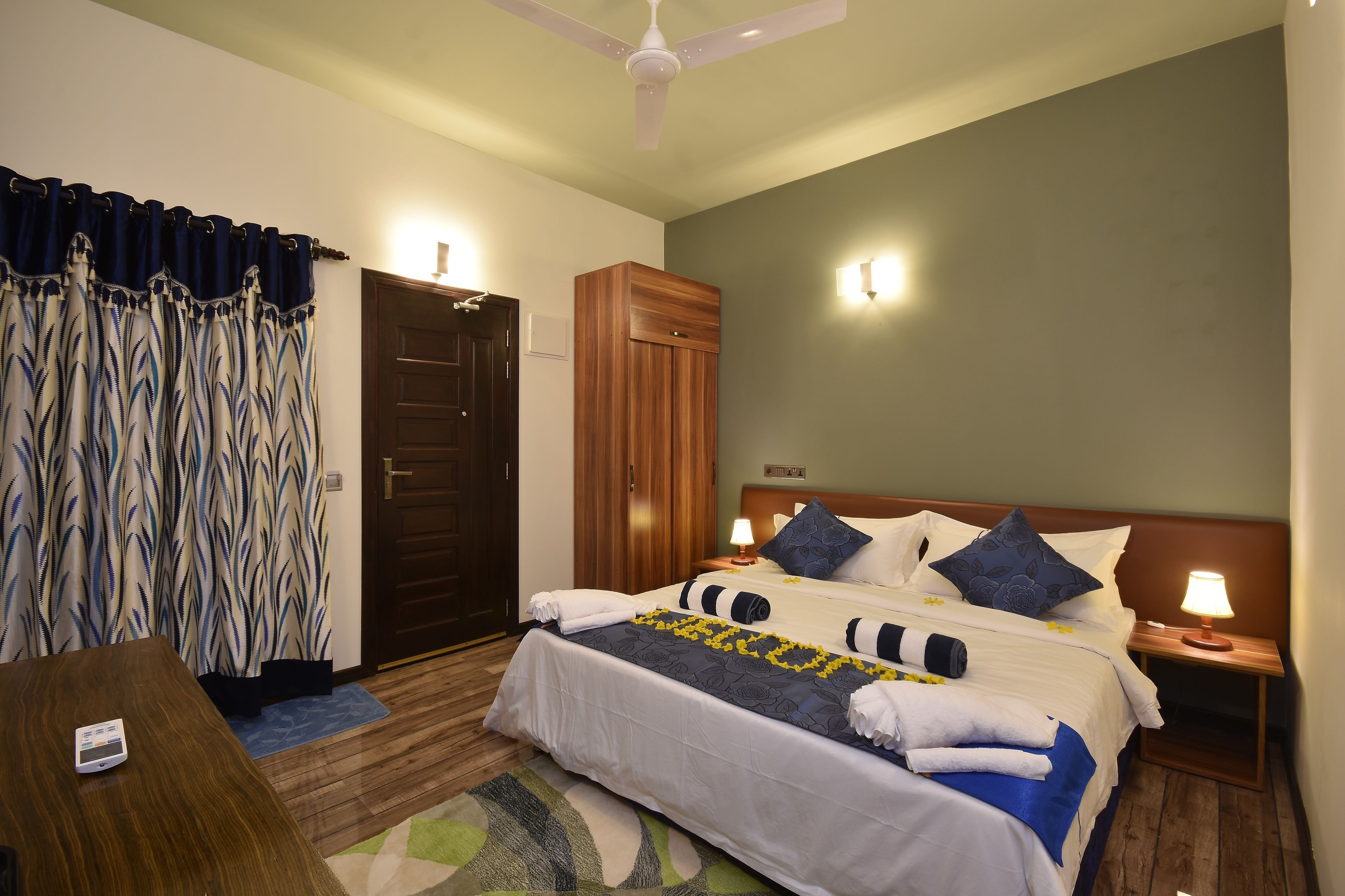 A hotel room with a bed, bedside tables, a desk, a chair, a wardrobe, and a ceiling fan.