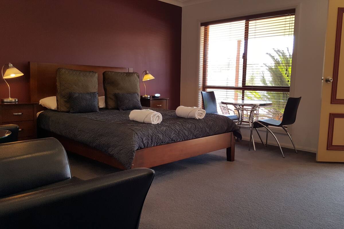A hotel room with a bed, two bedside lamps, a small table with chairs, and a window with blinds.
