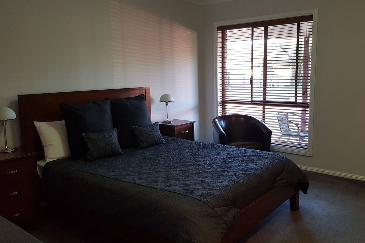 A bedroom with a large bed, a nightstand, a chair, and a window with blinds.