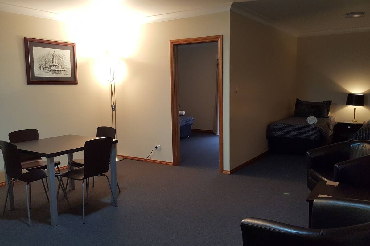 A hotel room with a dining table, chairs, a bed with a nightstand, a lamp, and a framed picture on the wall.