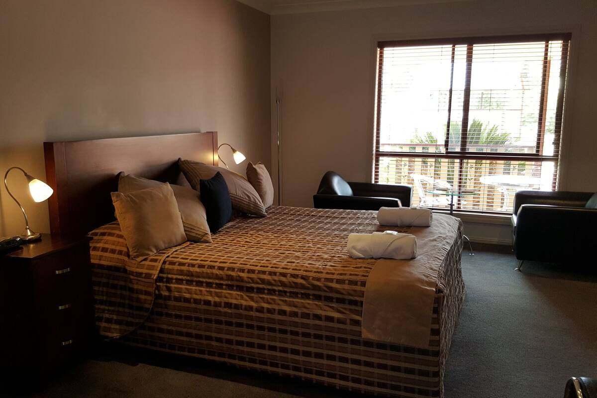 A bedroom with a bed, bedside lamp, chair, and a window with blinds.