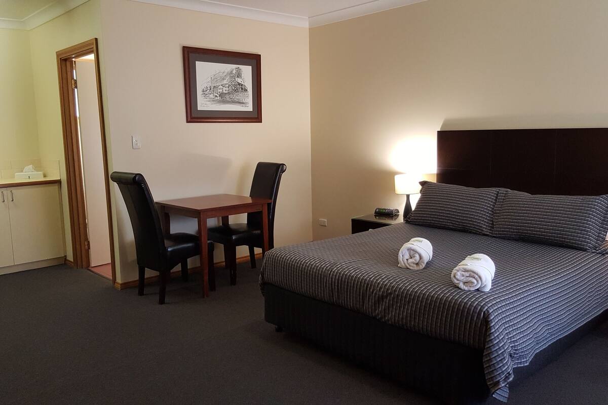 A hotel room with a bed, a desk, two chairs, a framed picture on the wall, and a nightstand with a lamp.