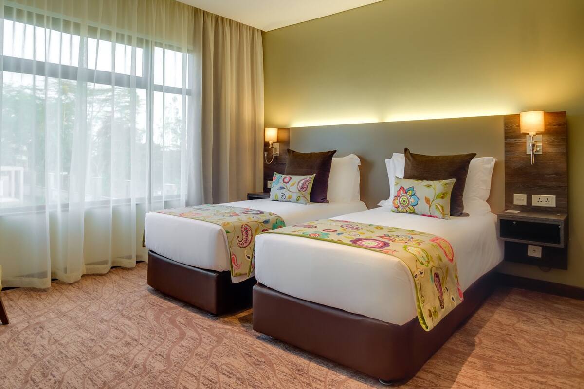 A hotel room with two beds, a chair, a nightstand, and a window with sheer curtains.