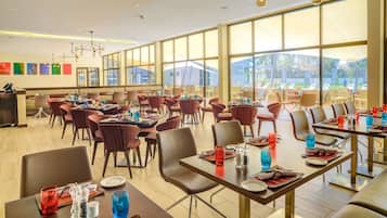 A spacious dining area with multiple tables and chairs, large windows offering views of the outdoors, and a modern chandelier.