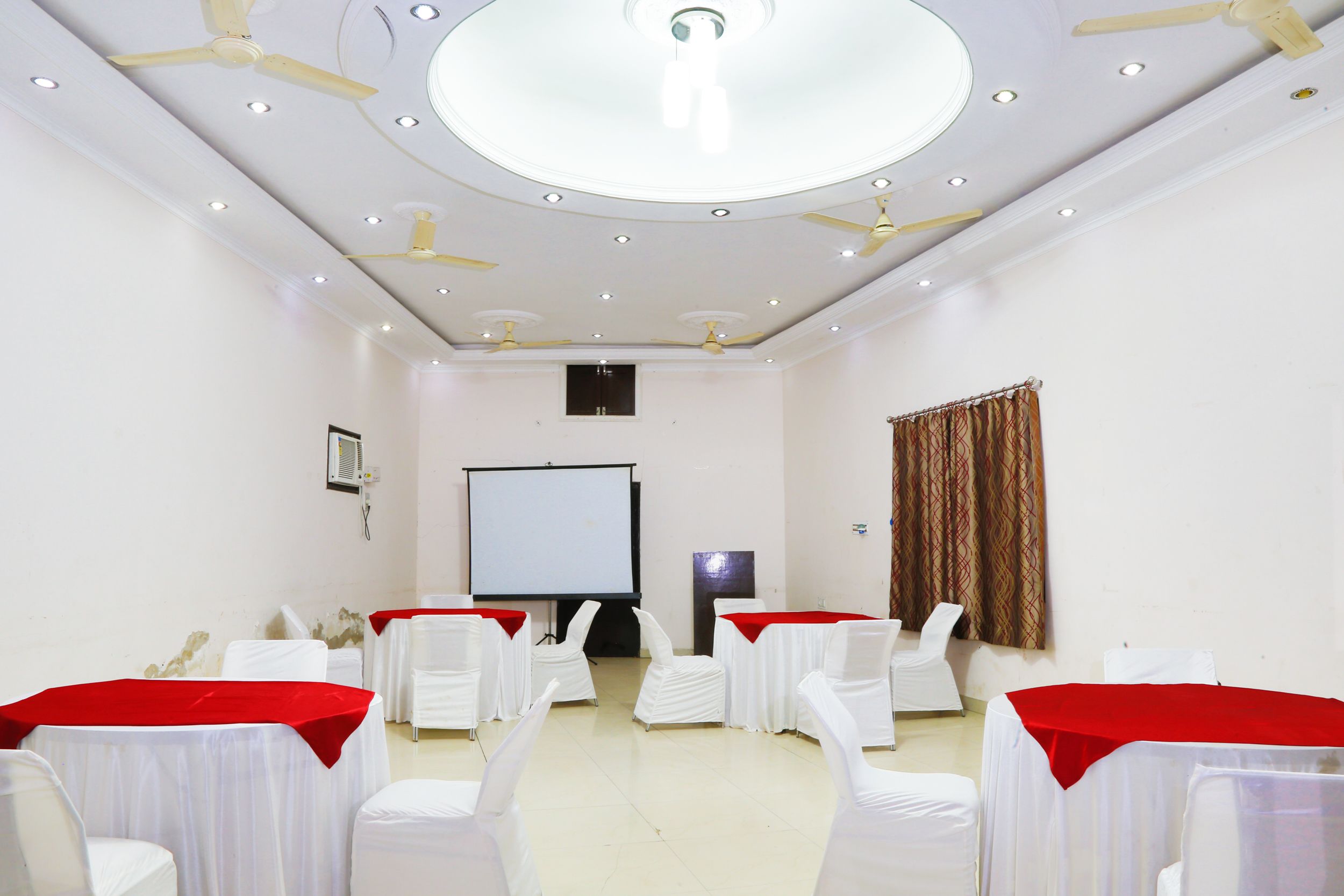 A conference room with white chairs, red tablecloths, ceiling fans, and a projector screen.