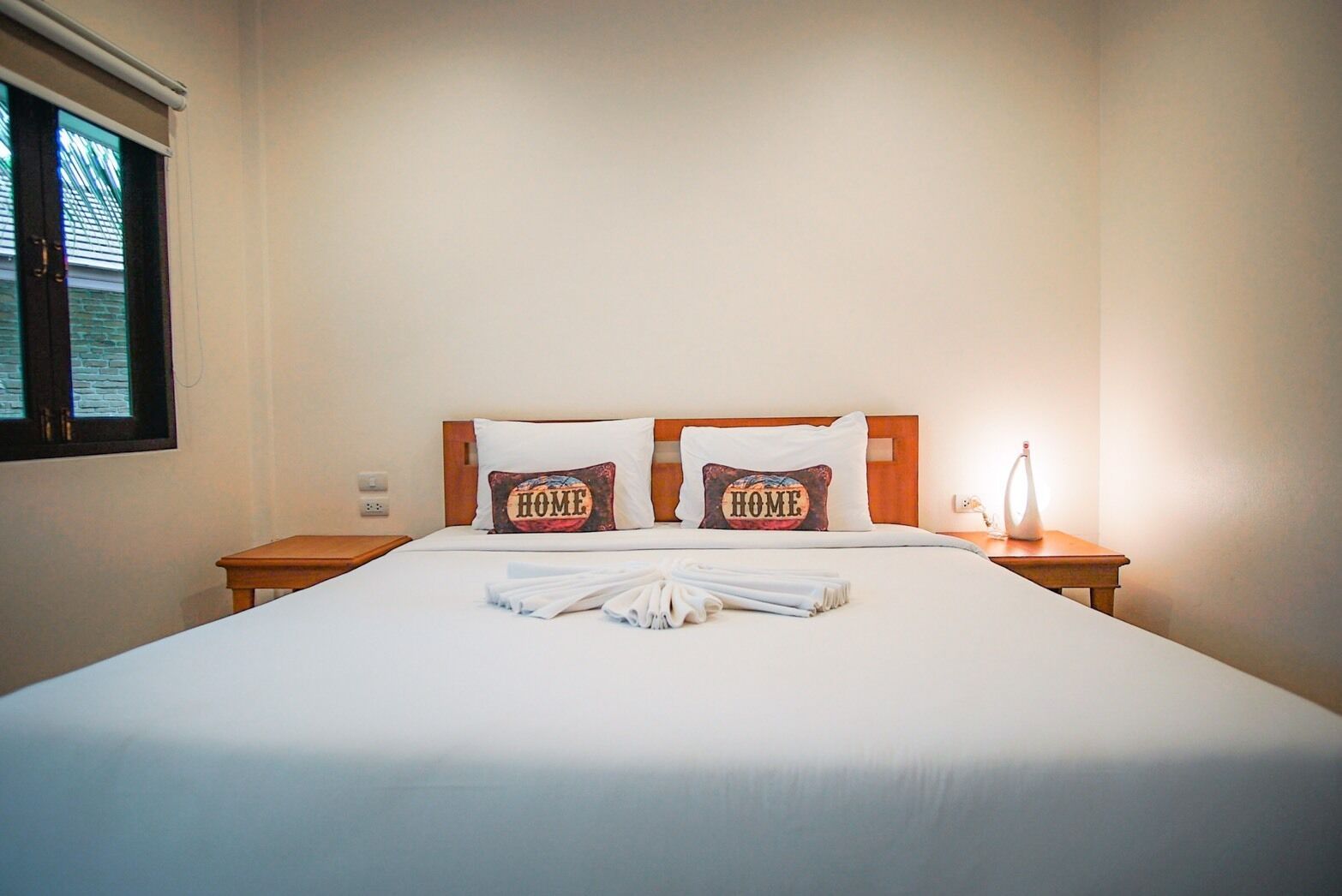 A neatly made bed with two pillows, a bedside table with a lamp, and a window with blinds.