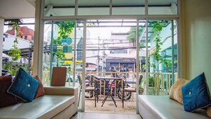 A room with a large glass door leading to an outdoor area with a table and chairs, a sofa with cushions, and a view of the street and buildings. - Capricorn Village (Patong)