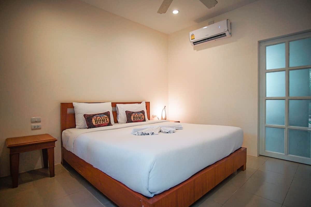 A hotel room with a large bed, two pillows with 'HOME' text, a bedside table, an air conditioner, and a window with frosted glass.