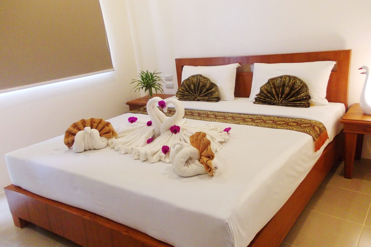 A hotel room with a neatly made bed, a wooden headboard, a bedside table with a lamp, and a potted plant.