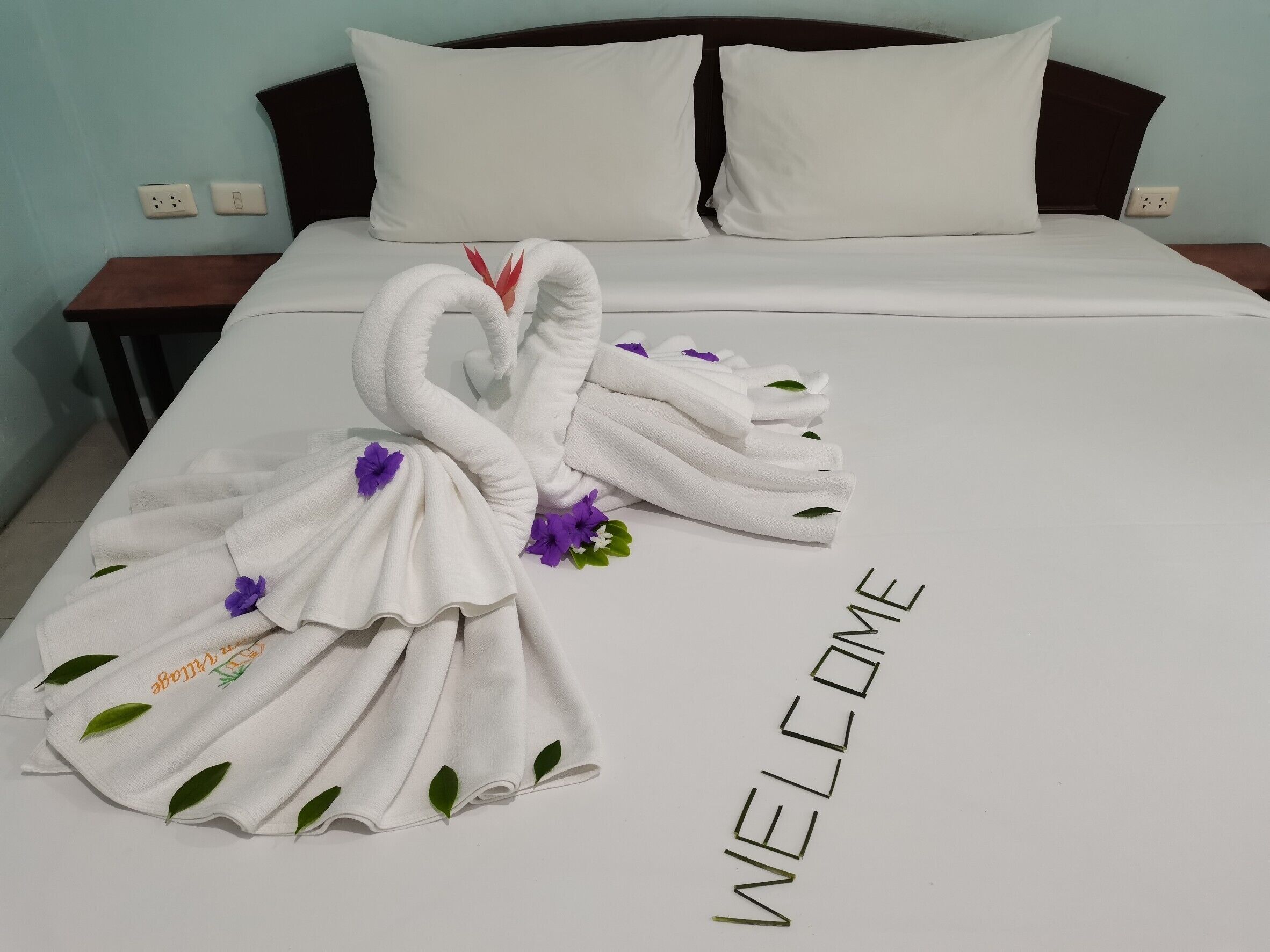 A neatly made bed with a towel swan and a 'WELCOME' sign, indicating a hotel room.