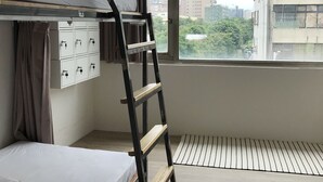 Um dormitório com beliches, uma janela com vista para edifícios e um radiador no chão.