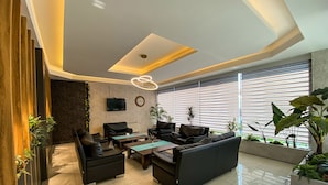 Una sala moderna con un sofá, sillones, una mesa de centro y un televisor montado en la pared. Hay plantas y un ventanal con persianas.