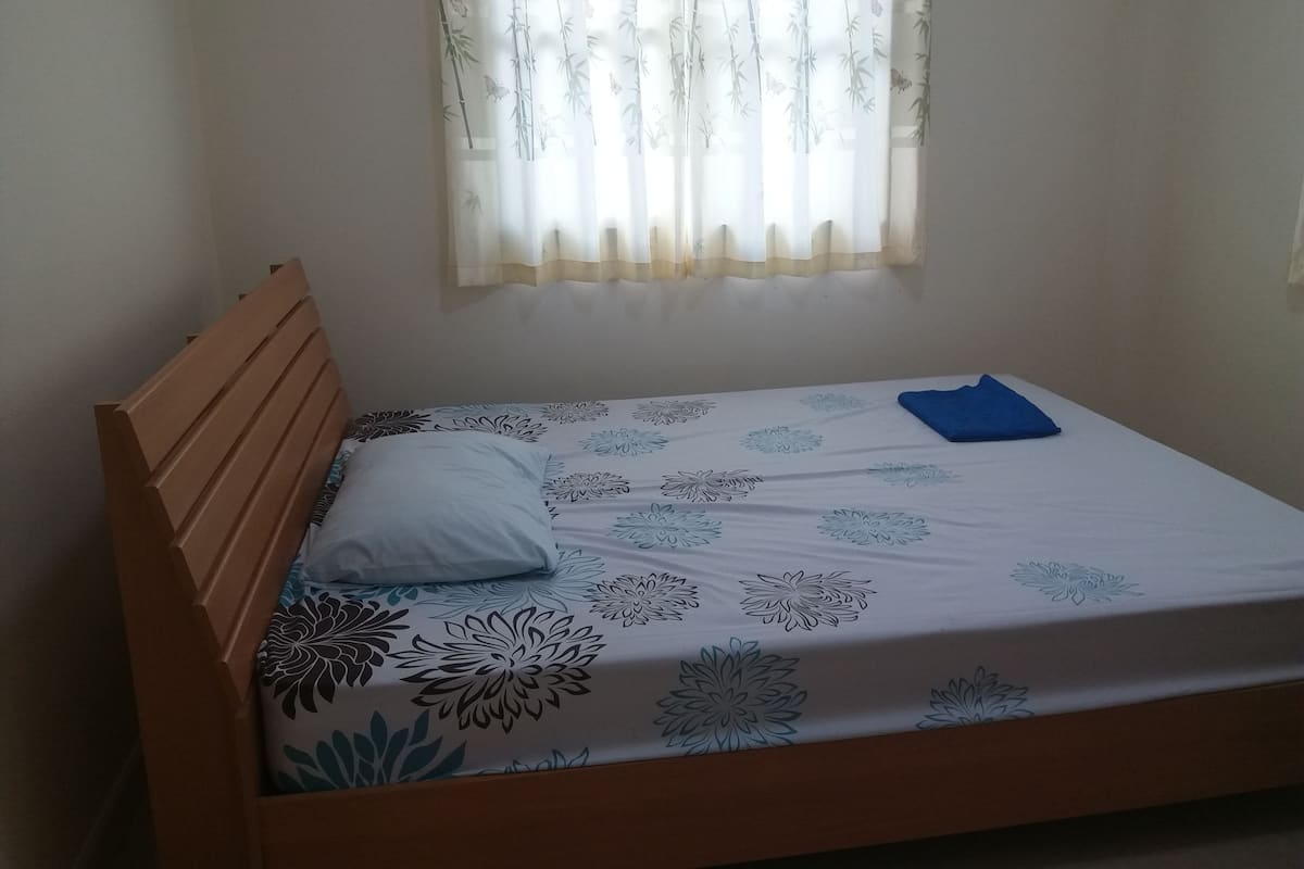 Uma cama de solteiro com cabeceira de madeira, colchão branco com estampas florais, travesseiro azul e uma janela com cortinas leves.