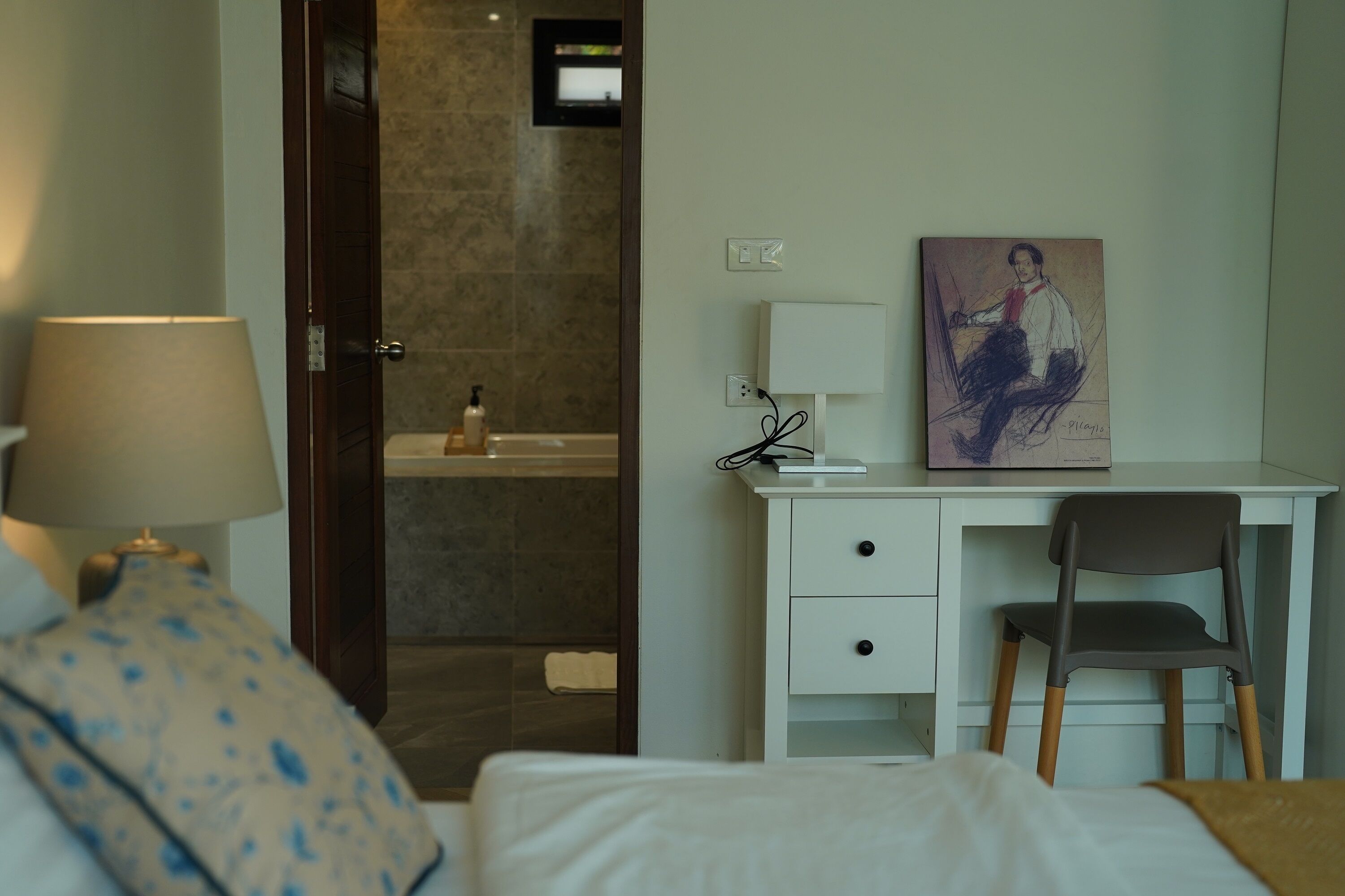 A hotel room with a bed, a desk with a chair, a lamp, a painting, and a bathroom visible through an open door.