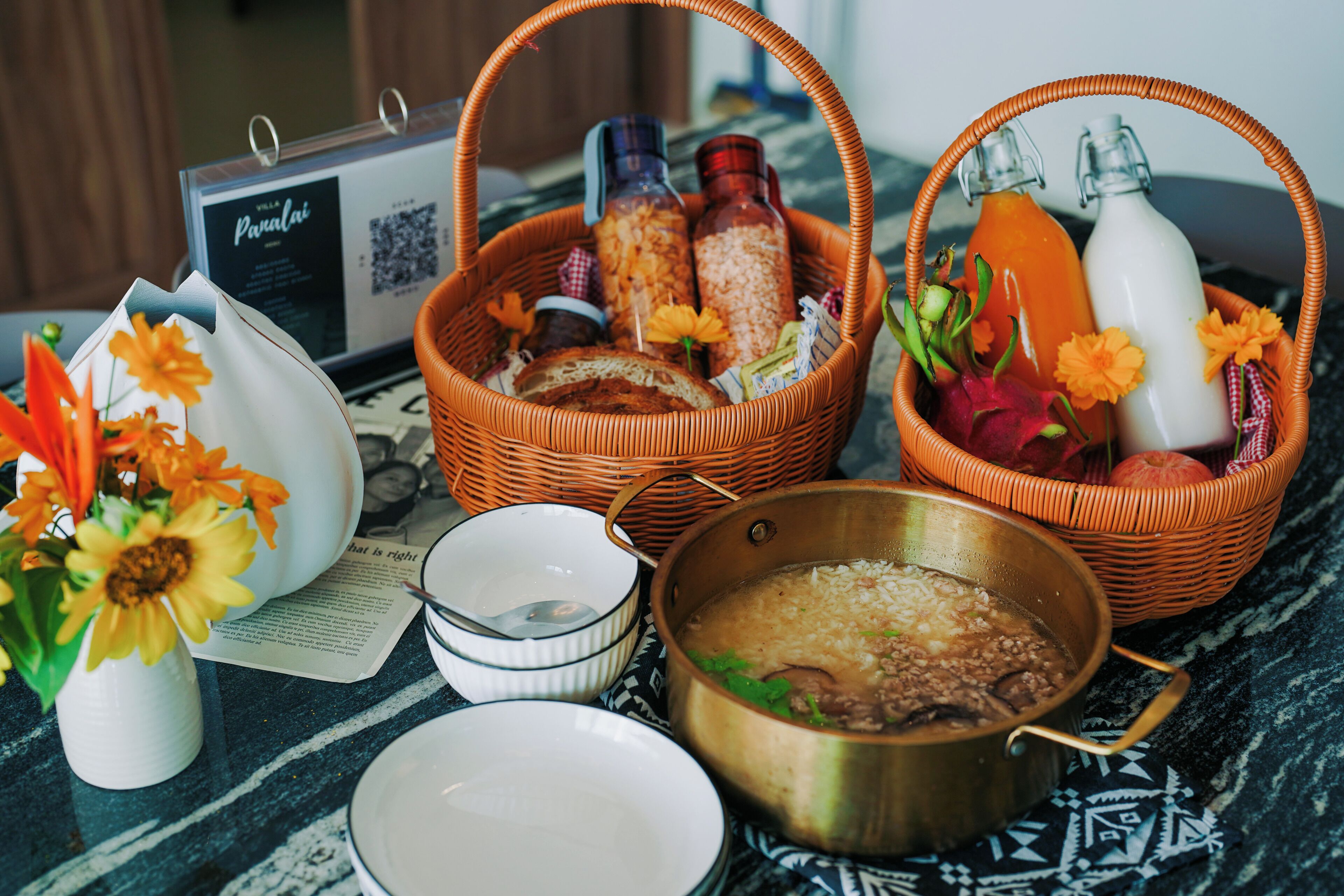 A table with a basket containing bottles, a bowl, a vase with flowers, and a pot with food.