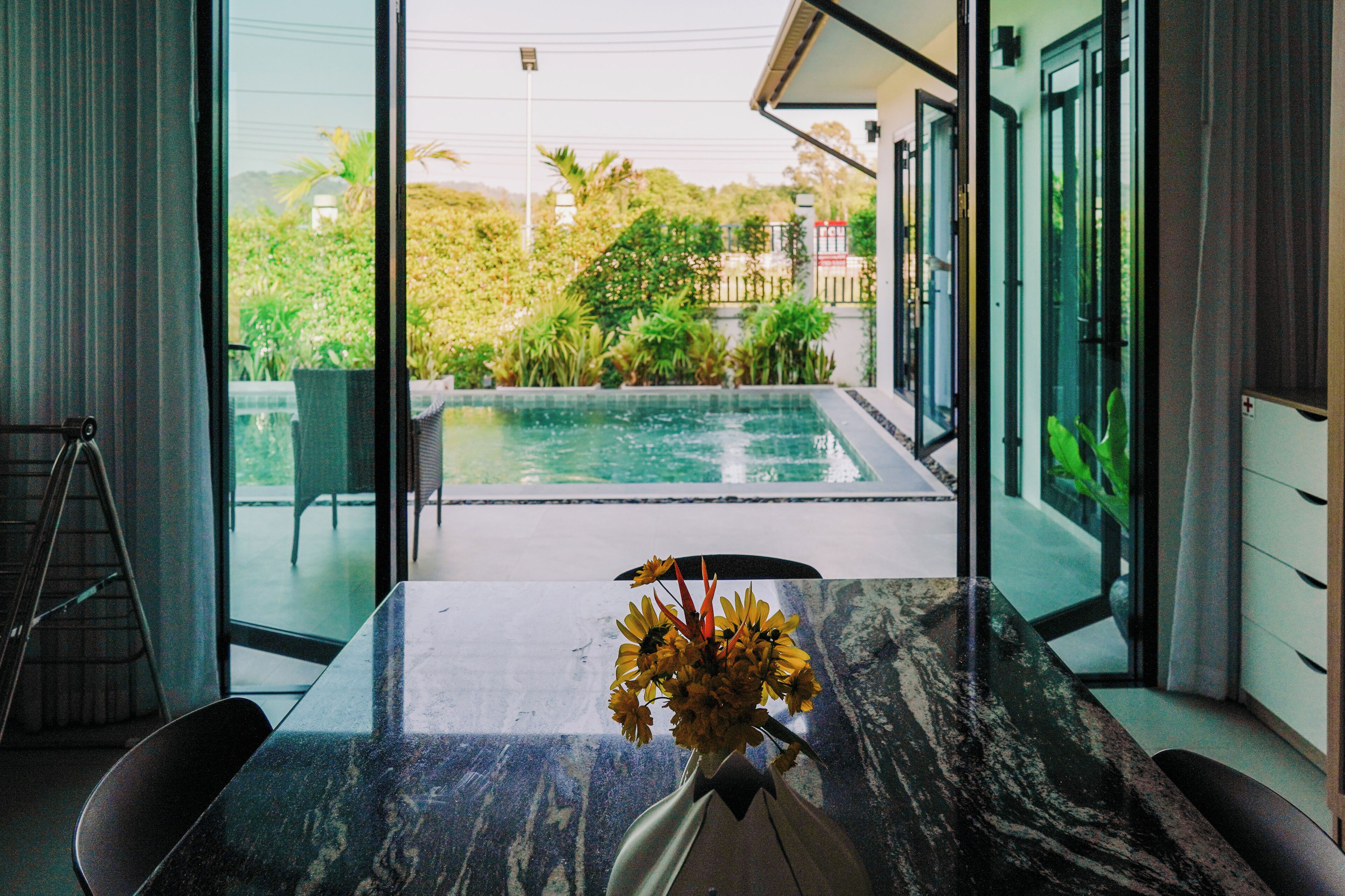 A modern interior with a marble table, a vase of flowers, and a view of a swimming pool and greenery outside.