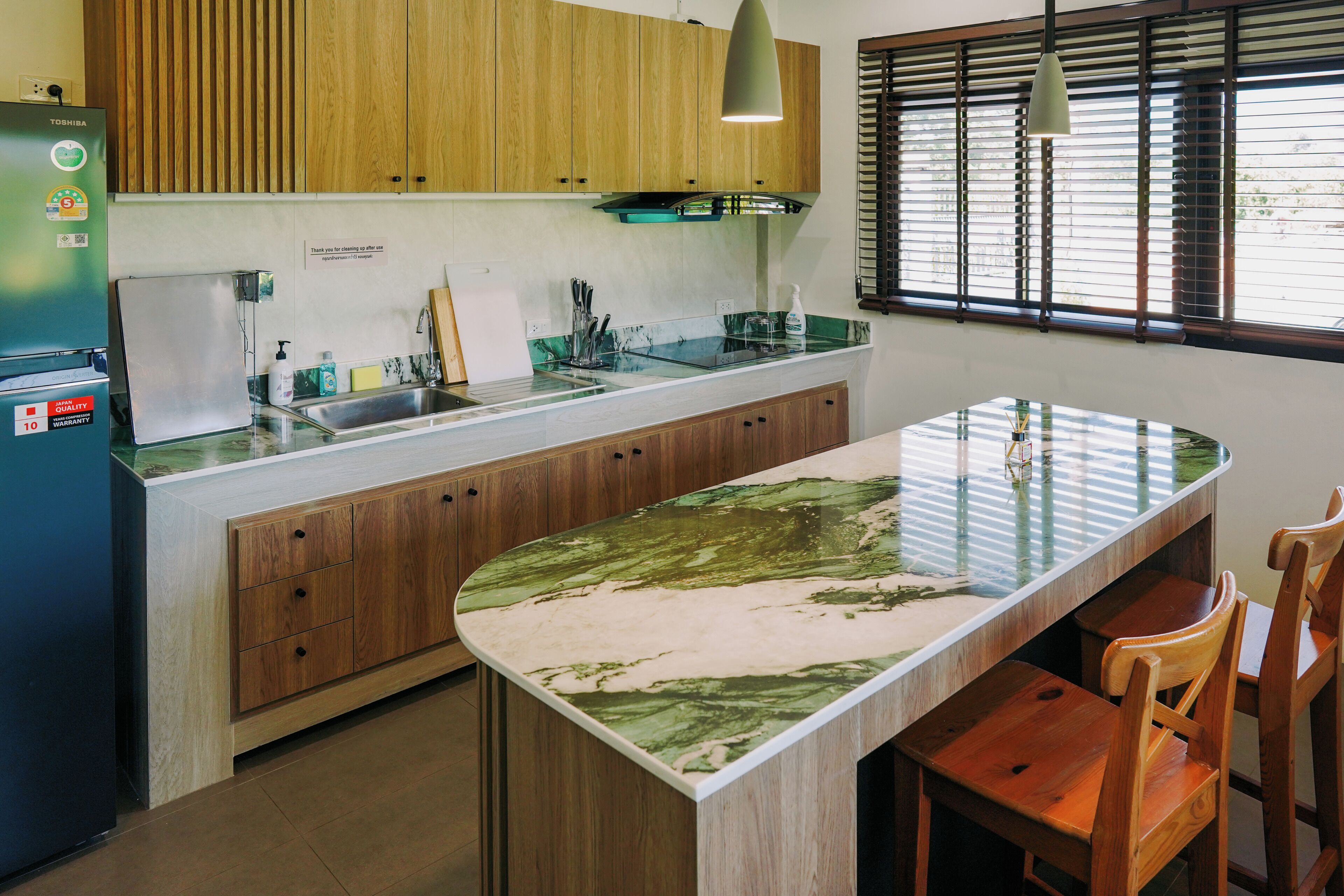 A kitchen with wooden cabinets, a built-in island, a refrigerator, and a window with blinds.