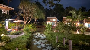 Een nachtzicht van een rivierengebied met een kleine beek, omringd door weelderige begroeiing en moderne gebouwen met verlichte ramen.