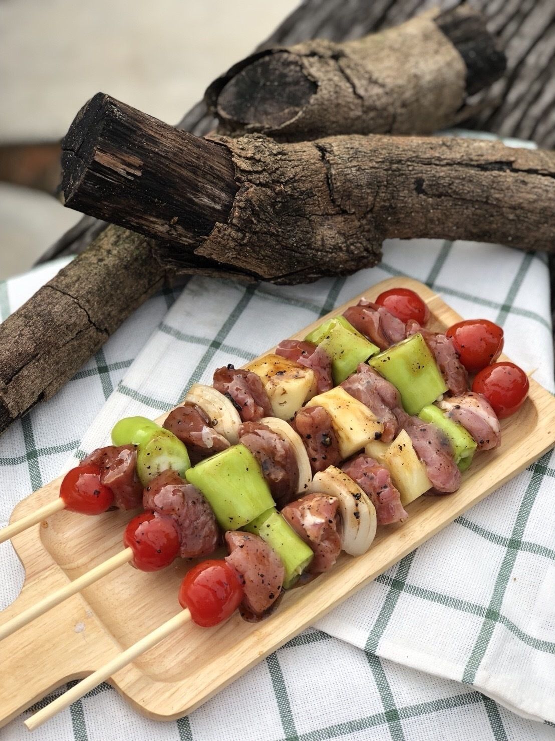 A wooden skewer with meat and vegetables, placed on a checkered cloth with a piece of driftwood in the background.