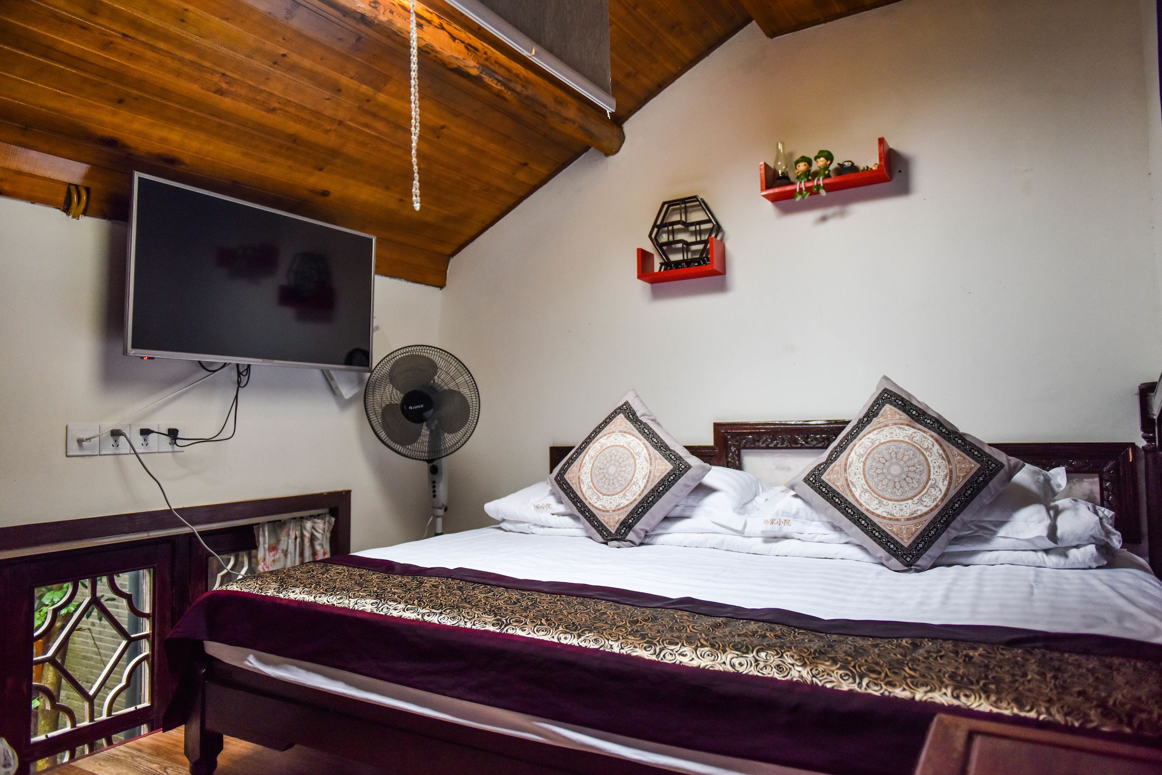 A bedroom with a bed, a TV mounted on the wall, a fan, and a window with a decorative view.