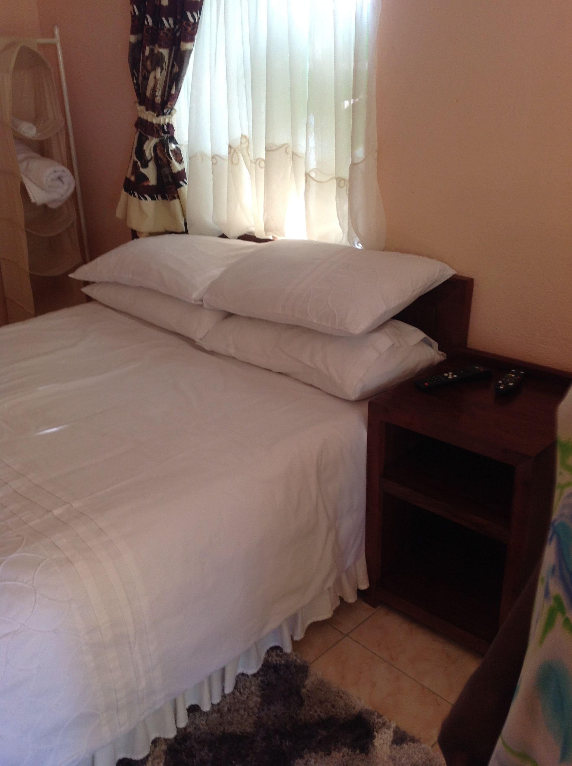 Una cama bien tendida con ropa blanca, una mesita de noche de madera con un control remoto y una ventana con cortinas de gasa.