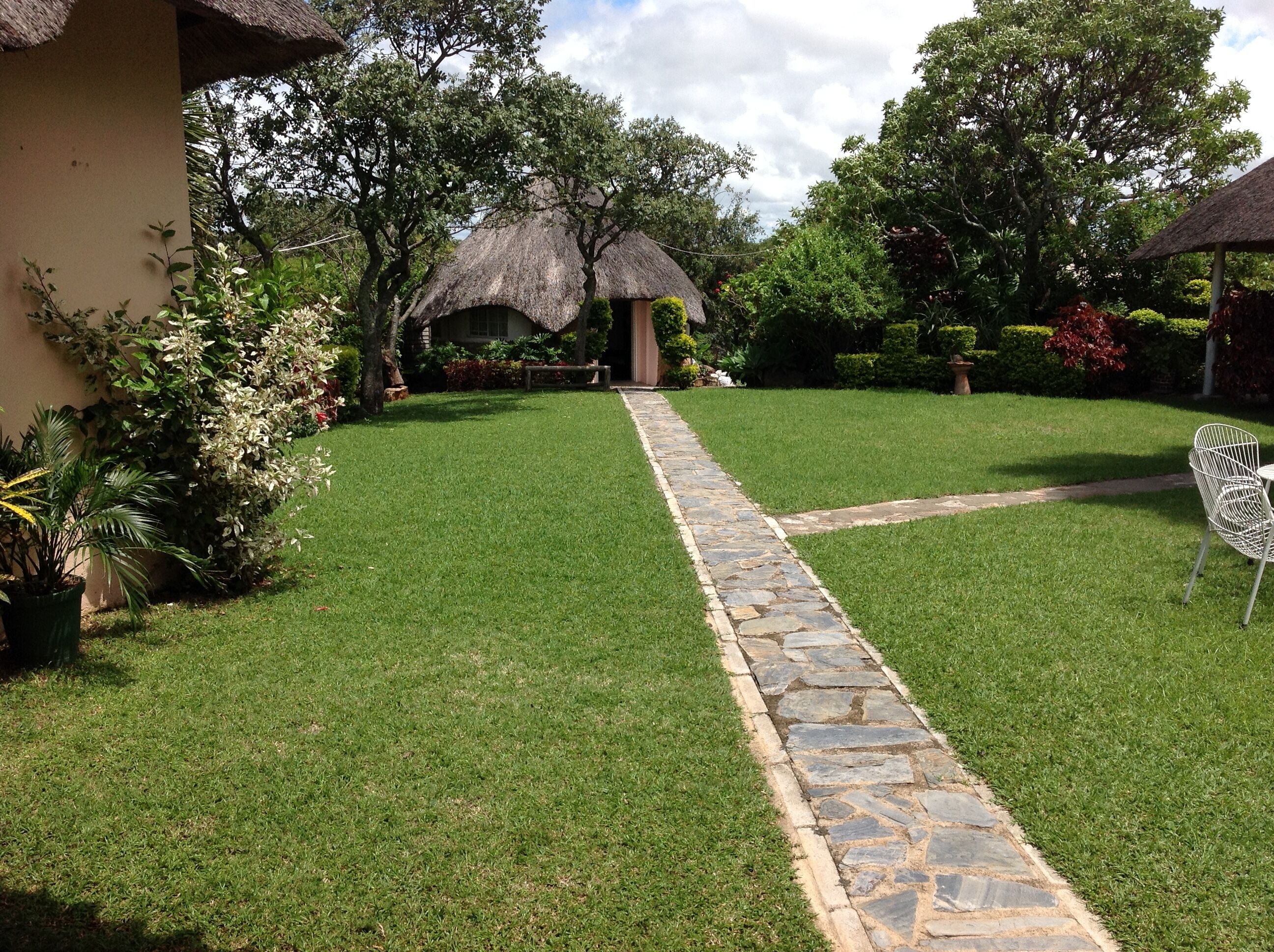 A well-maintained garden with a stone pathway leading to a thatched-roof structure, surrounded by lush greenery and potted plants.