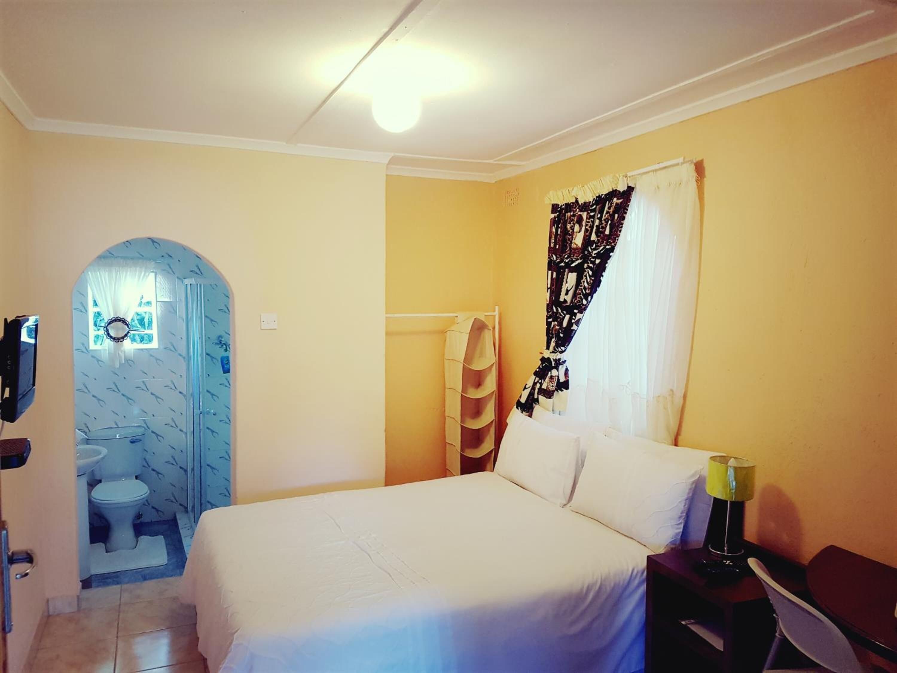 Un dormitorio con cama, mesita de noche, lámpara, ventana con cortinas, zona de ducha y baño con inodoro y lavabo.