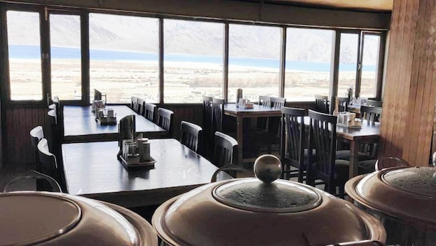 A restaurant with large windows offering a view of a snowy landscape. Tables are set with chairs and covered with tablecloths. - TIH Ladakh Summer Camp Pangong (Leh)