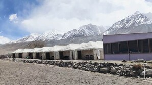 A row of white tents with a mountain backdrop, a stone wall, and a modern building with large windows. - TIH Ladakh Summer Camp Pangong (Leh)