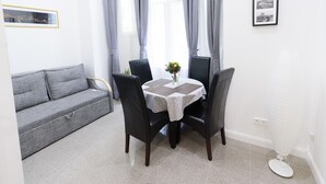 Un petit salon, aménagé avec soin, comprenant un canapé gris, une table ronde avec des chaises et un petit vase avec des fleurs.