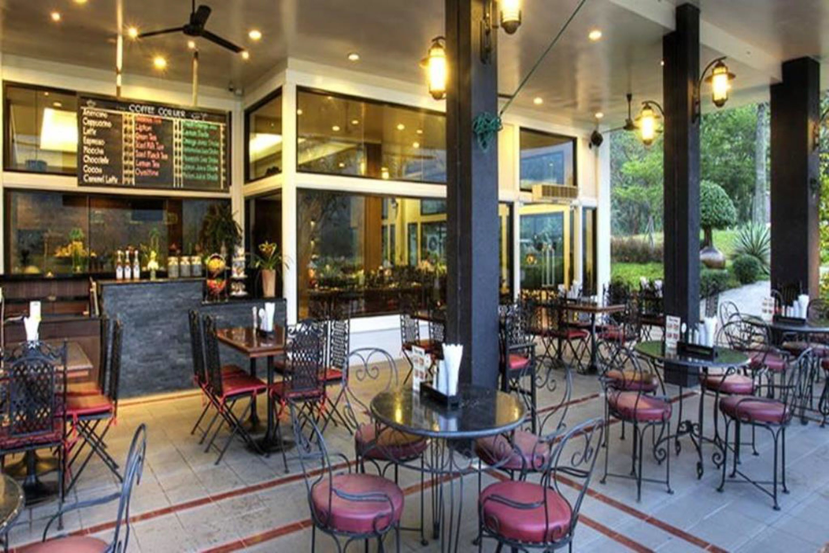 A spacious outdoor dining area with round tables and chairs, a coffee bar with a menu, and large windows offering views of the surrounding greenery.