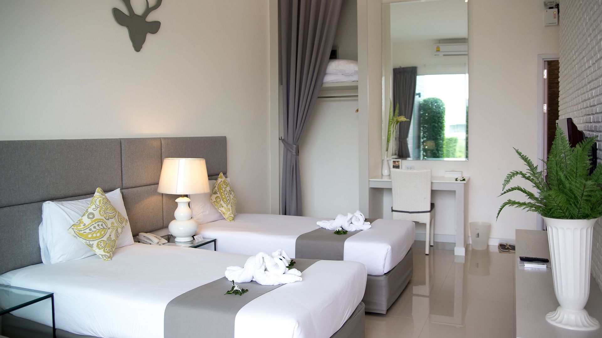 A hotel room with a bed, bedside tables, a lamp, a mirror, a desk, and a potted plant.