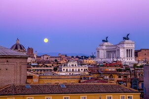 Paesaggio urbano con edifici storici, un imponente monumento bianco e una luna piena nel cielo.