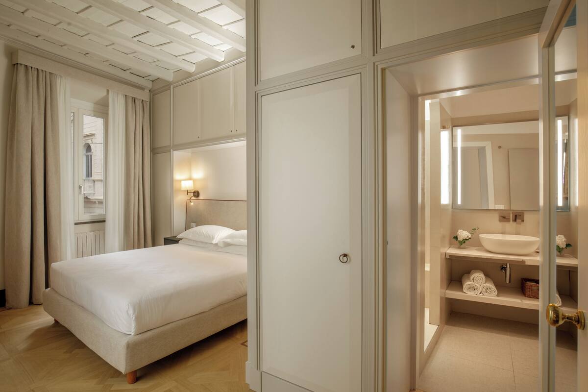 A bedroom with a bed, a nightstand, a wall-mounted lamp, and a bathroom with a sink and mirror.