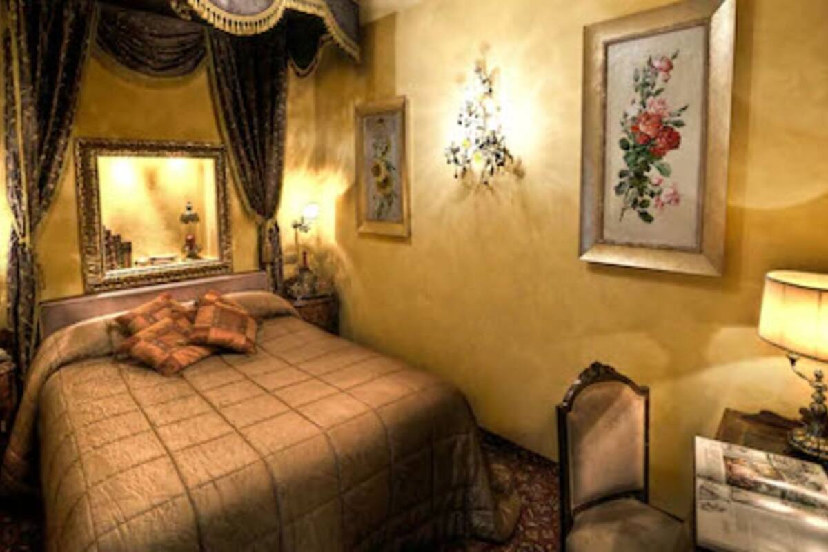 A hotel room with a large bed, a nightstand, a chair, a desk, a lamp, a mirror, and a floral painting on the wall.