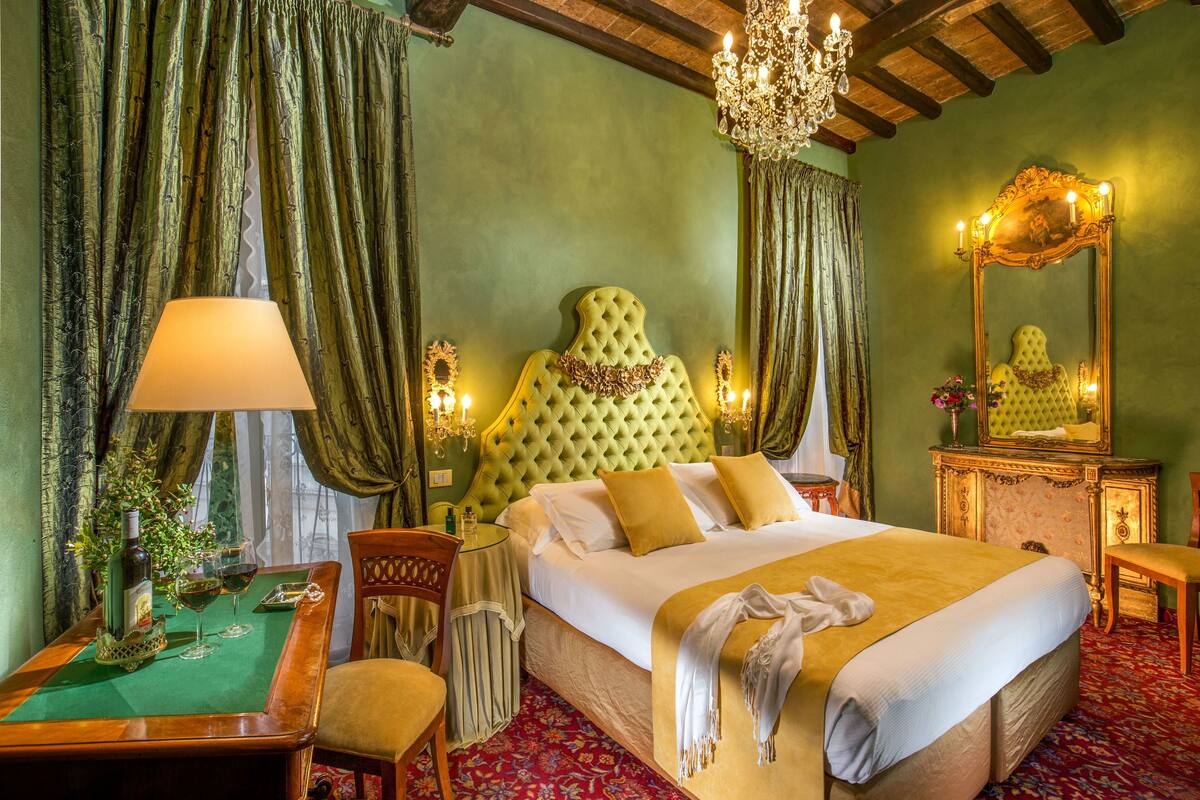 A hotel room with a large bed, a chandelier, a mirror, and a table with a bottle and glasses.