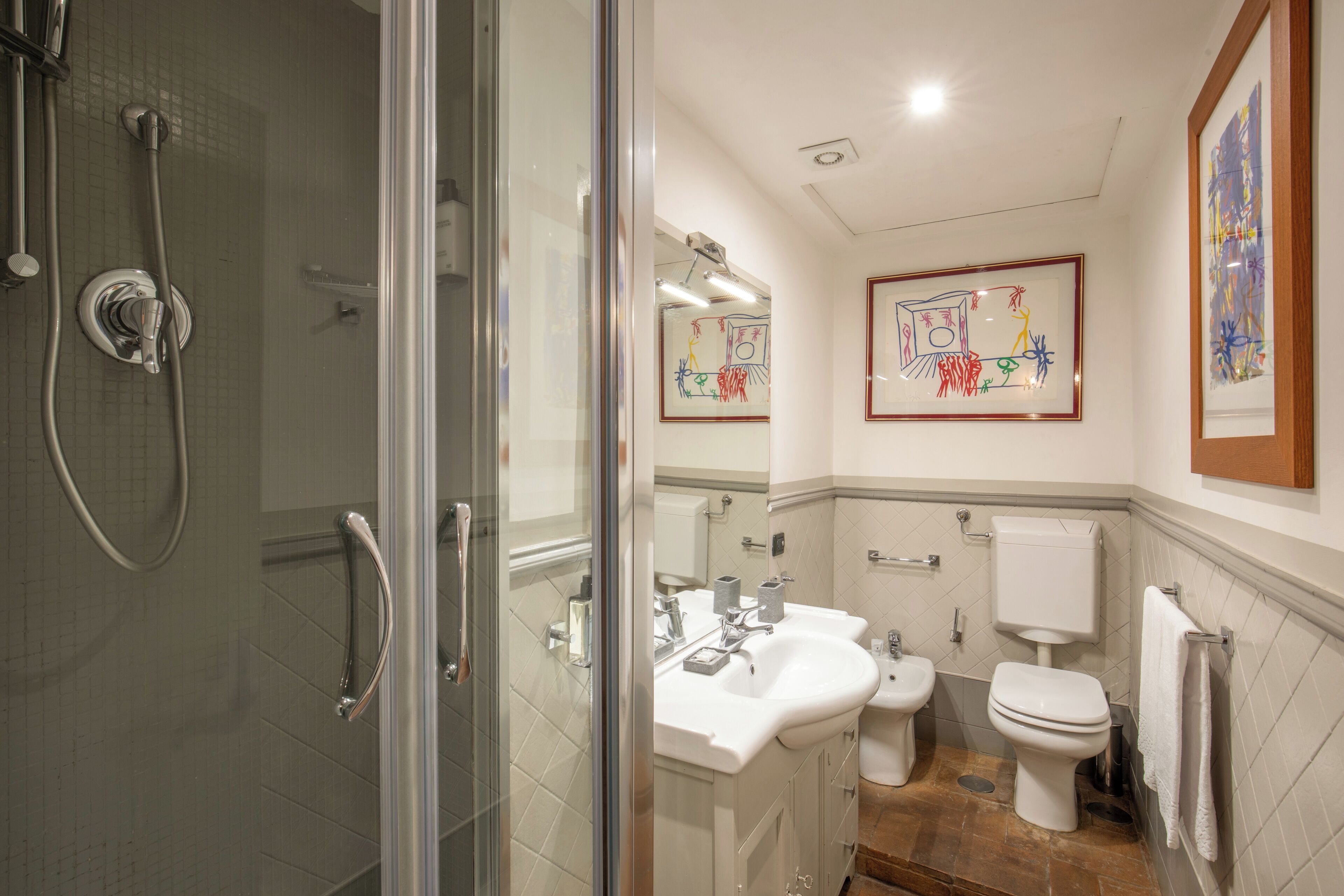 A modern bathroom with a glass shower enclosure, a double sink vanity, a toilet, and framed artwork on the walls.