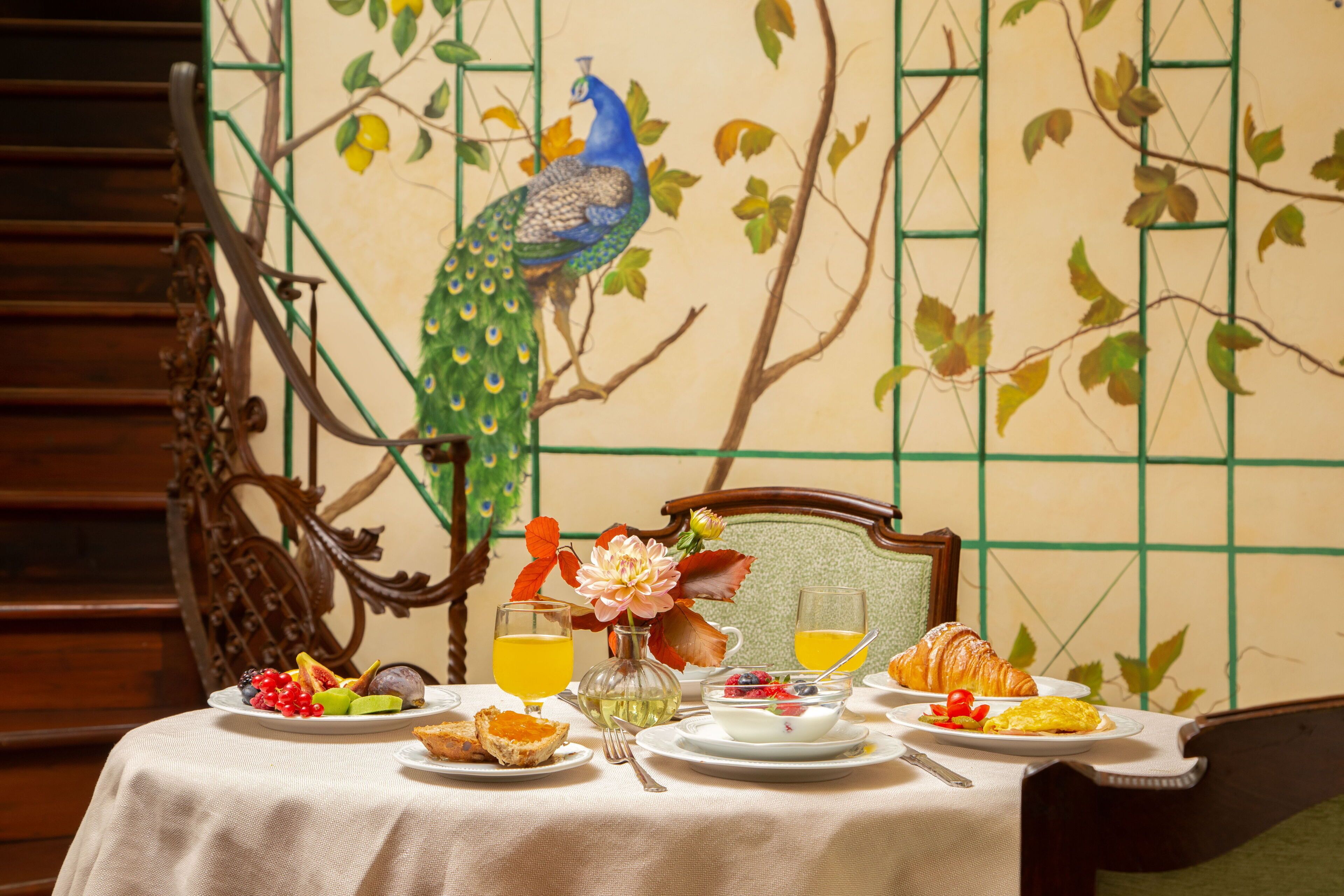 Una mesa de desayuno con platos de comida, un florero con flores y un mural de un pavo real en la pared.