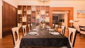 A dining area with a long table set for six, surrounded by wooden chairs. A chandelier hangs from the ceiling, and a wooden cabinet with glass doors displays various bottles and decorative items. - Villa Xilaiwan (Ratsada)