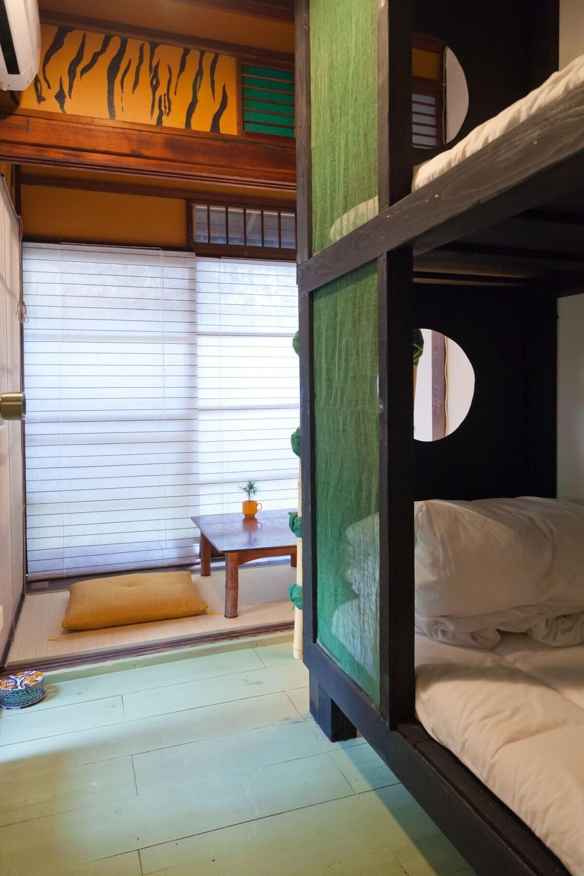 Ein Zimmer mit einem Etagenbett, einem kleinen Tisch mit Pflanze und einem Fenster mit Jalousien.