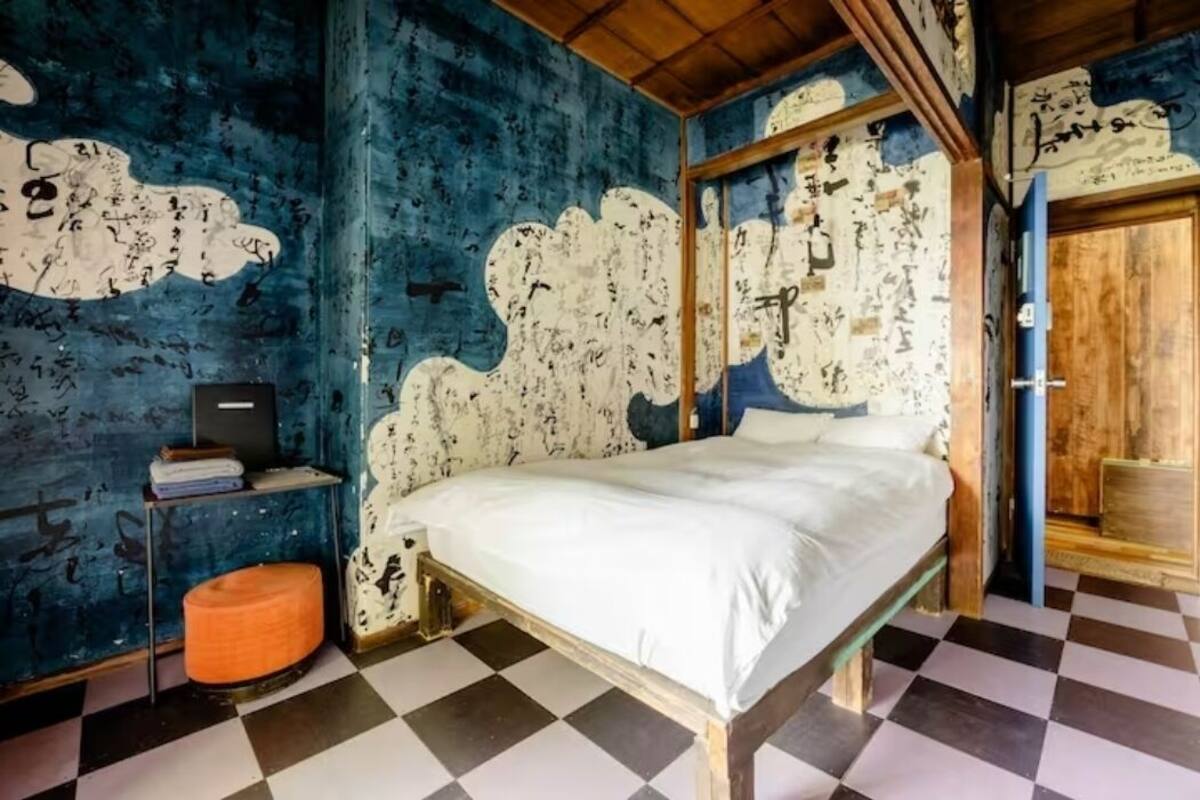 A bedroom with a bed, a small table, and a chair. The walls have a unique pattern, and the floor has a checkered design.