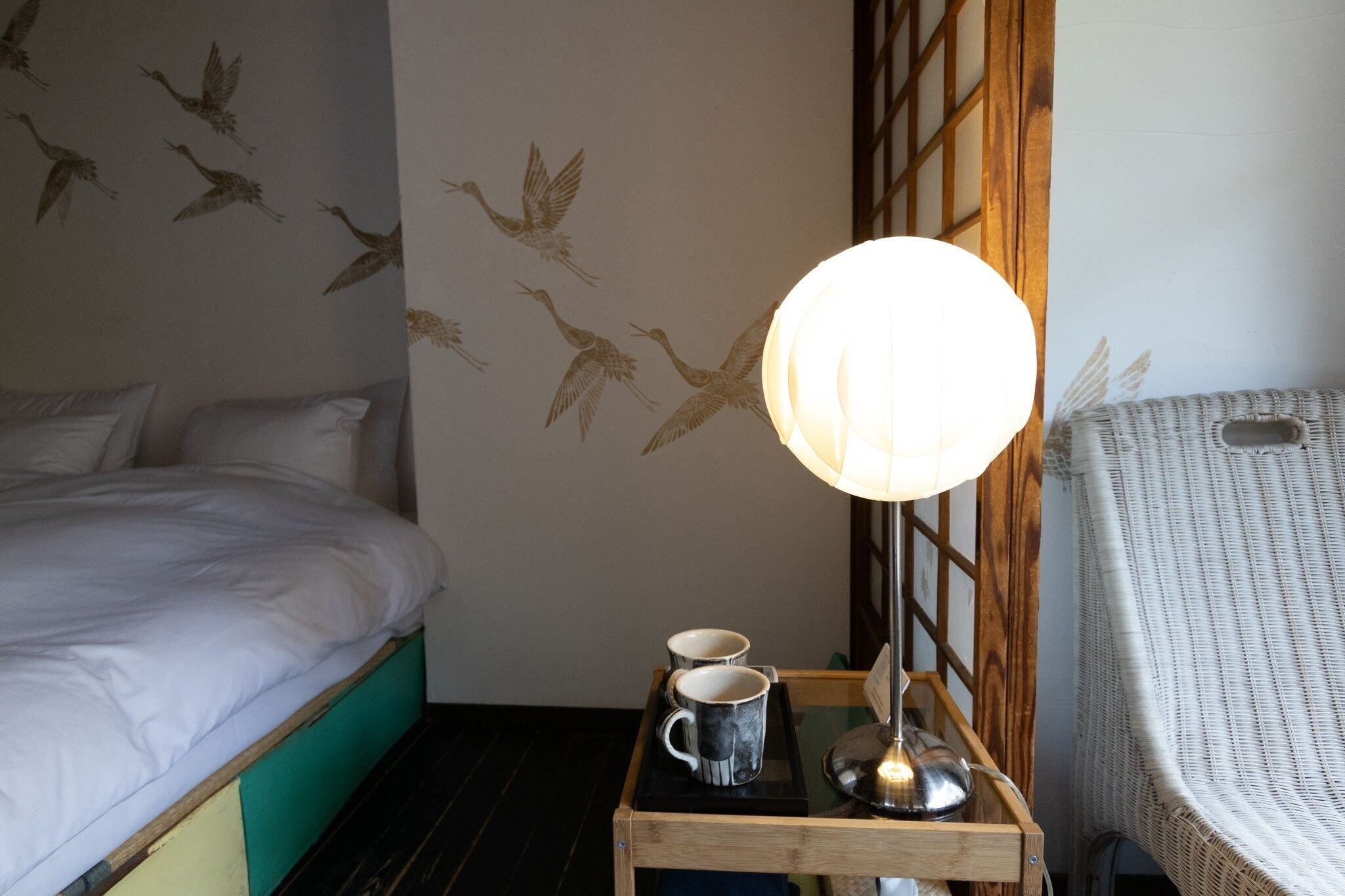 A hotel room with a bed, a bedside table with two mugs, a lamp, and a wall with a bird mural.