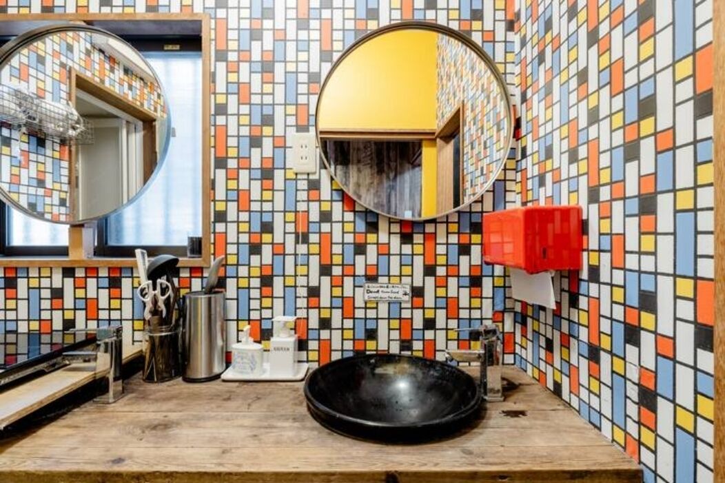 Een badkamer met een wand betegeld met kleurrijk mozaïek, een ronde spiegel, een wastafel en een rode zeepdispenser.