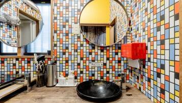 Een badkamer met een wand betegeld met kleurrijk mozaïek, een ronde spiegel, een wastafel en een rode zeepdispenser.