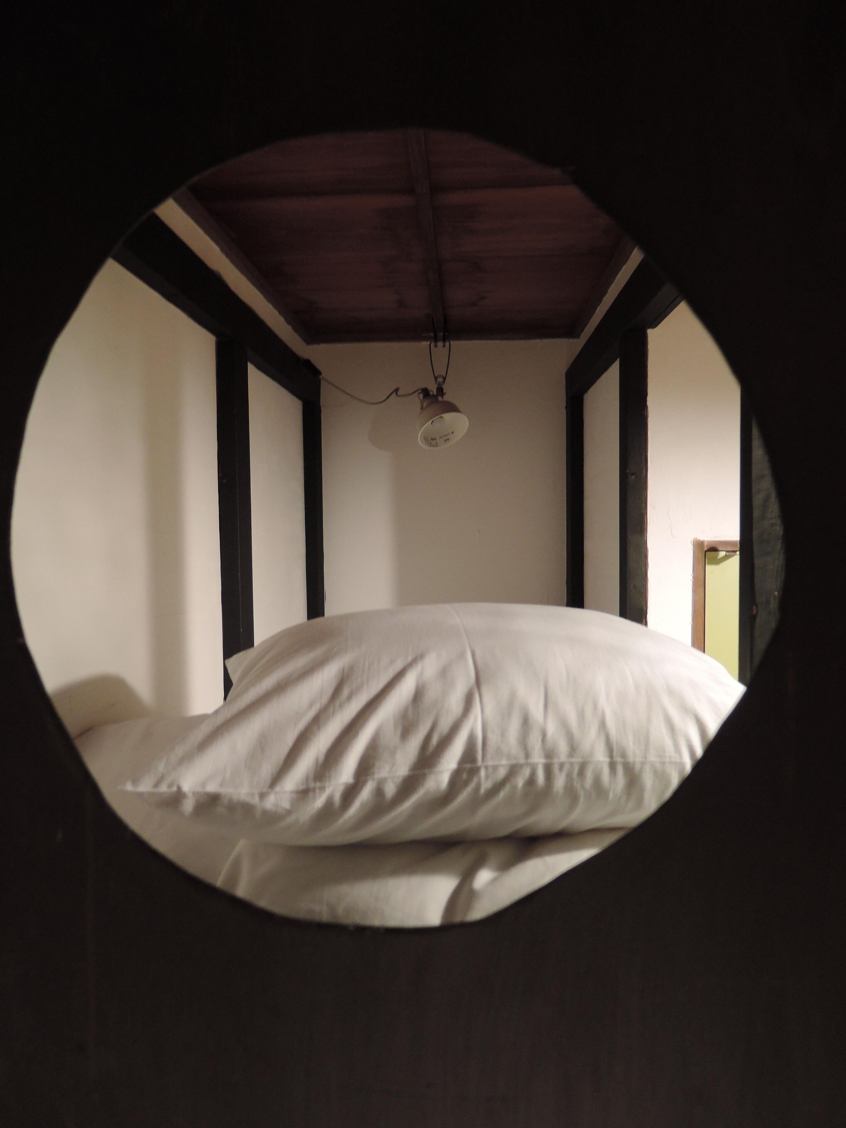 Runder Blick durch ein Fenster auf ein Schlafzimmer mit Bett, Holzbalken und Deckenventilator.