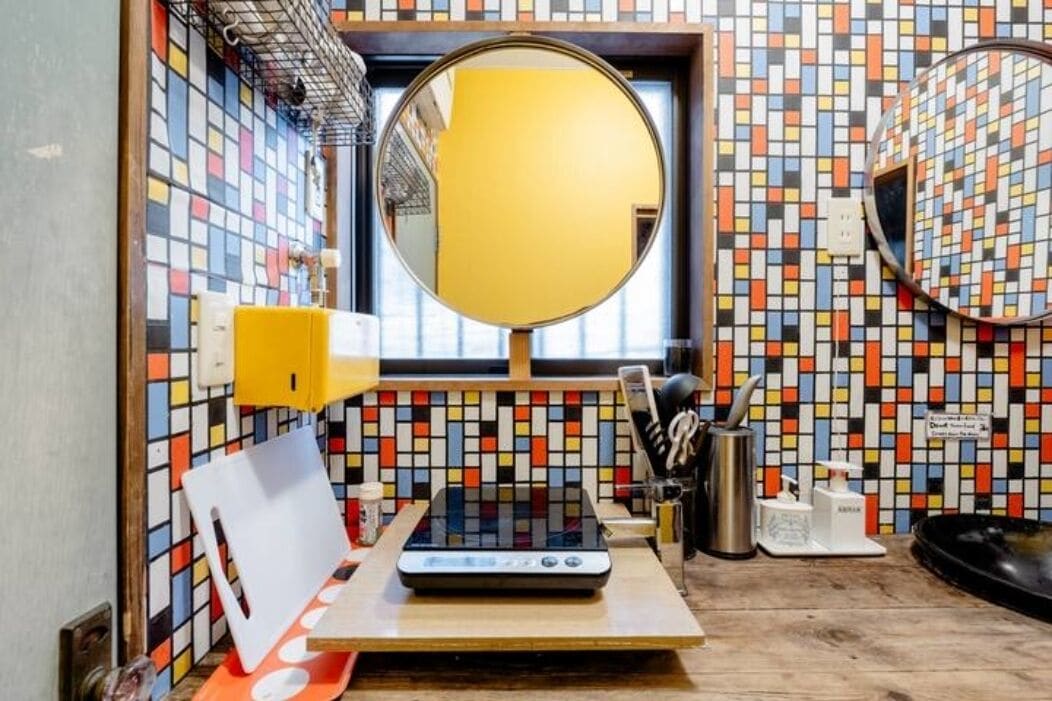 A bathroom with a colorful mosaic tile backsplash, a round mirror, a sink with a yellow soap dispenser, and a small table with a scale and a white object.
