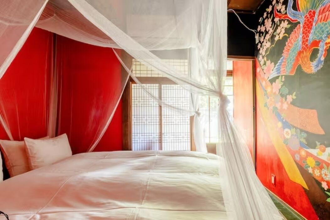 A room with a bed, red walls, a window with blinds, and a mural with a colorful design.