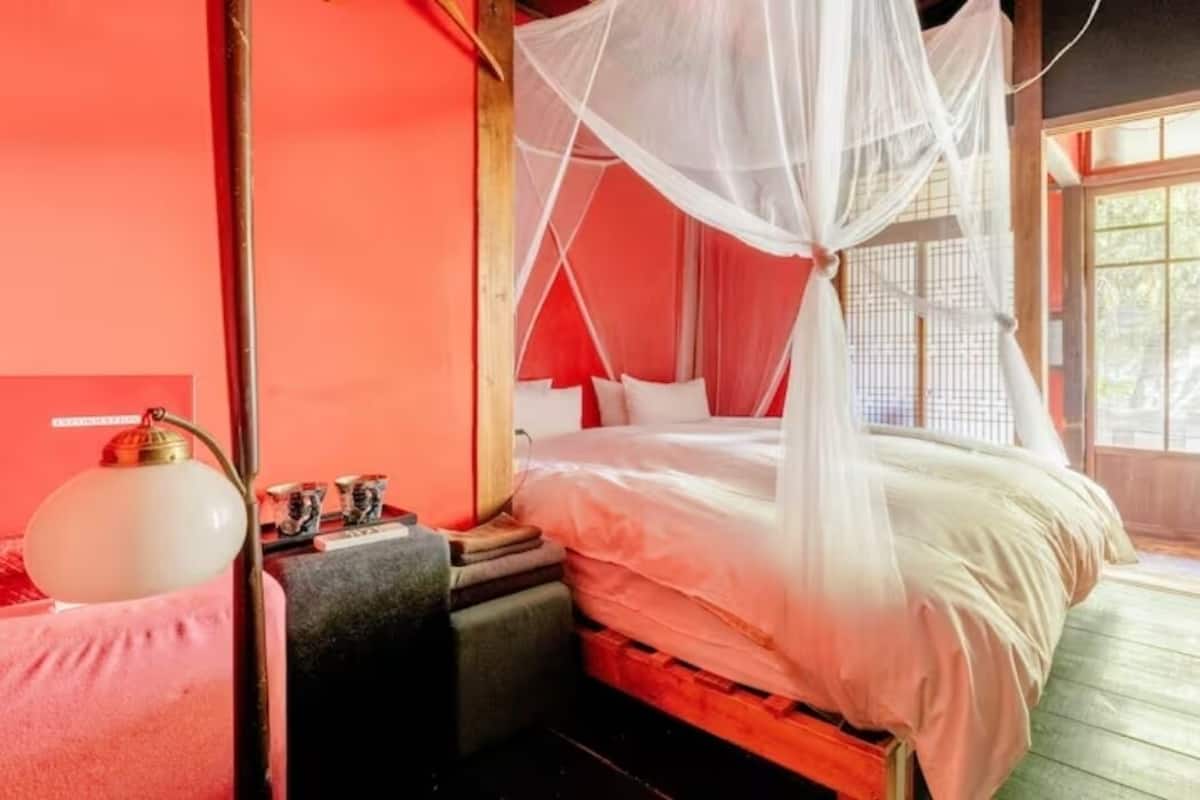 A room with a bed under a white canopy, a small table with a lamp, and a window with a view.
