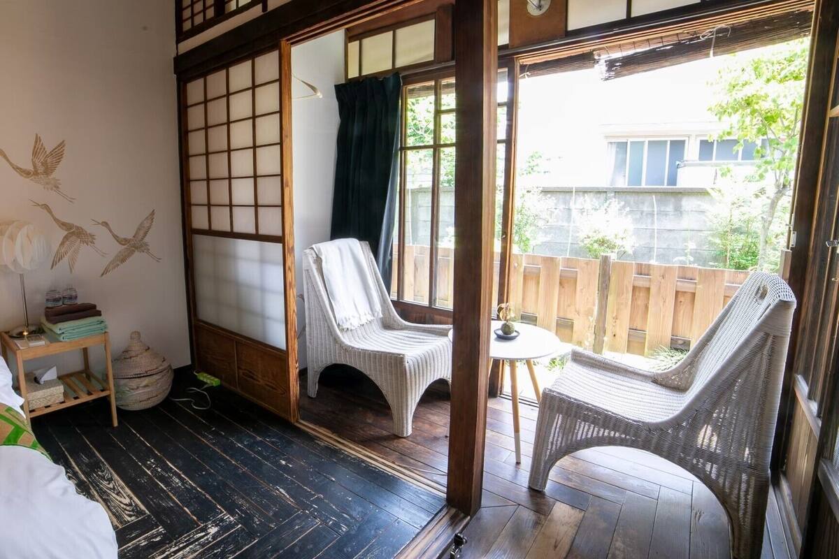 A traditional Japanese room with tatami flooring, a sliding shoji screen, a bed with a green patterned blanket, and a view of the outside.