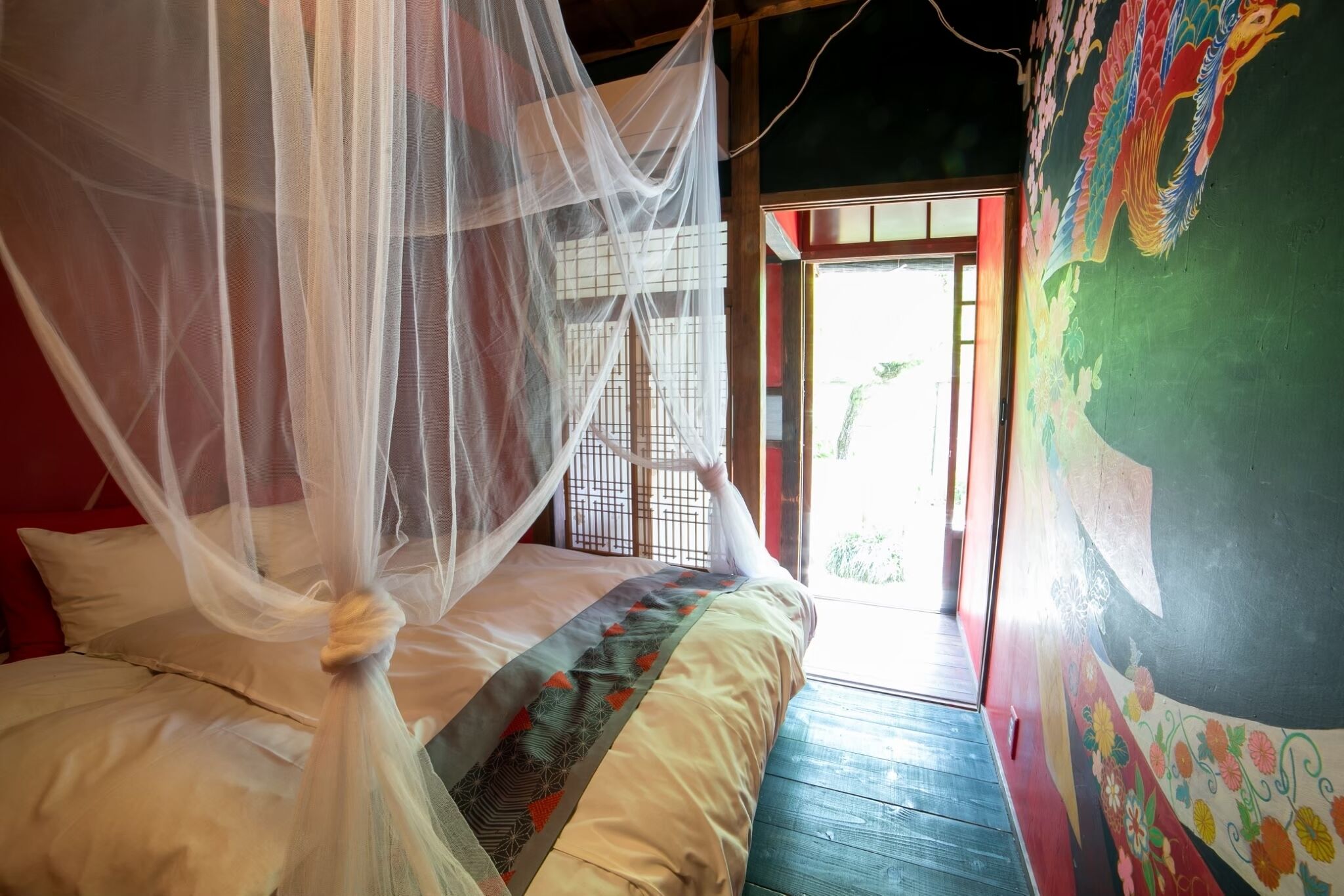 A room with a bed covered by a mosquito net, a wooden floor, and a colorful mural on the wall.